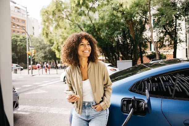 Smiling traveler leaning on her electric car while it charges