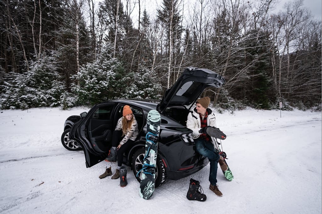 Couple with snowboards getting ready beside their electric car in the snow